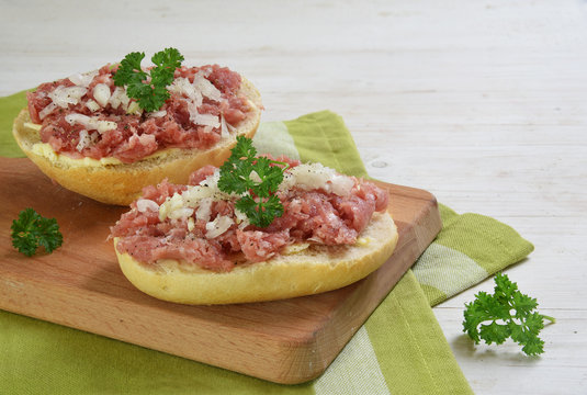 Bread rolls with minced pork sausage, german zwiebelmettwurst with onions and parsley garnish on a breakfast board,  green napkin and white wooden background with copy space
