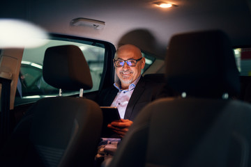 Fototapeta na wymiar Mature professional happy successful businessman is being driven in the back seat of the car while using a tablet.