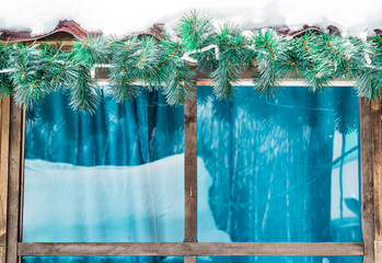 Wooden house window decorated under the roof with artificial fir tree branch with icicles and snow, winter Christmas background
