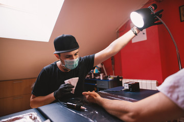 Young professional Tattoo artist in black cloths and sterile mask on adjusting light toward...