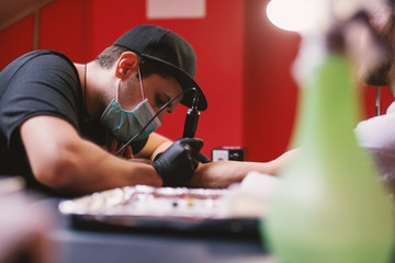 Young focused tattoo artist is inking customers arm carefully in his shop.