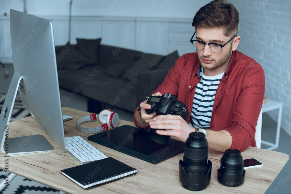Fototapete Man freelancer with camera in hands at work by table with computer and graphic tablet