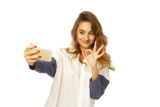 Portrait of a beautiful young woman making selfie on smart phone and hand ok sign. Happy woman photographing herself.