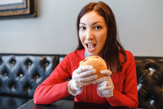 Happy young woman eating a hamburger at indoor cafe in protective white gloves