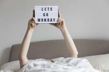 Female in bed under the sheets holding up a lets go monday sign