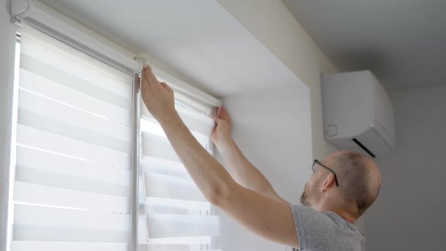 adult man is installing a rod for blinds on a window frame in a living room in daytime and fixing curtains