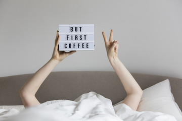 Female in bed under the sheets holding up a but first coffee sign