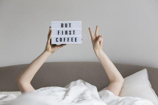 Female In Bed Under The Sheets Holding Up A But First Coffee Sign