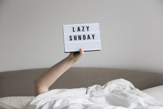 Female in bed under the sheets holding up a lazy sunday sign