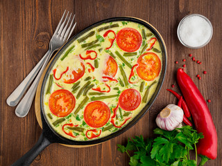 Oval cast iron pan with delicious hot omelette with tomatoes and capsicum. Brown wood texture background, close-up, top view.