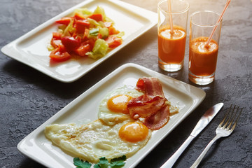 Breakfast from fried egg, bacon, vegetable salad and carrot juice. On a dark stone background