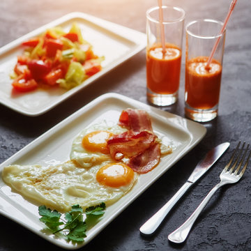 Breakfast from fried egg, bacon, vegetable salad and carrot juice. On a dark stone background