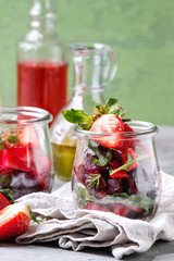 Beetroot and strawberry salad served with arugula and nuts in glass jars with cloth, pepper and bottles of fruit ocet and olive oil over grey table with green wall as background. Healthy eating