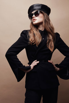 Fashion portrait of beautiful young woman in black flared jacket, black leather beret cap and dark sunglasses with veil. Standing with hands on waist