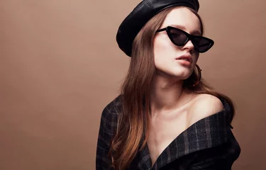 Fashion portrait of beautiful young woman in black leather beret cap, plaid jacket and cat eye retro sunglasses with massive golden earrings © monchak