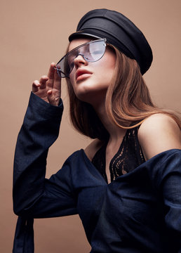 Fashion portrait of beautiful young woman in black leather beret cap, denim blouse and blue retro sunglasses