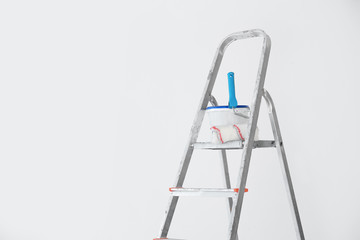 Bucket with paint and roller on step ladder near white wall