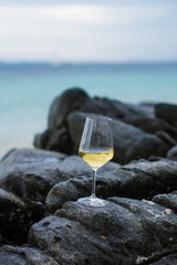 White wine glass on the rock in natural view on the beach of Thailand. Travel on vacation concept.  Glass of white wine on black stones coast in seascape background.