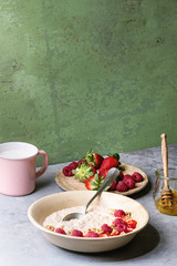 Sweet rice porridge pudding in ceramic plate with berries strawberry and raspberry, walnuts, honey and mug of milk on grey kitchen table with green wall as background.