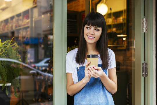 Smiling woman standing at entrance door of a store holding takeaway coffee
