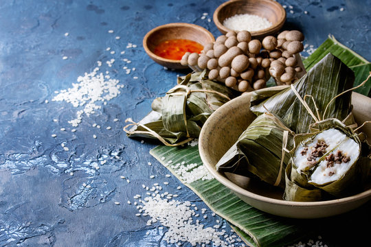 Asian rice piramidal steamed dumplings from rice tapioca flour with meat filling in banana leaves served in ceramic bowl. Ingredients and sauces above over blue texture background. Close up, space.