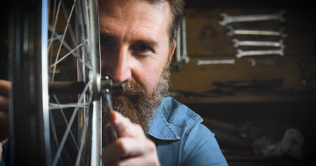 Well-groomed handsome bearded master hipster, specialist in bicycles, repairing a bicycle in his workshop, wheels, frame, spokes, the background of tools. Concept: pro bike, cycle passion, lifestyle.