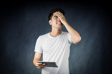 Terrible headache. portrait handsome young asian man wearing a white t-shirt holding smart phone or tablet  feeling stressful isolated on black background. Businessman concept. Asia people.