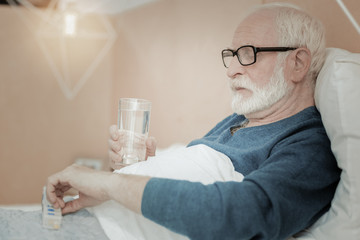 Time to treatment. Senior ill bespectacled man lying on the bed holding a glass of water and taking pills.
