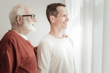 Good weather. Satisfied pleasant gay men standing in the bright room smiling and looking through the window.