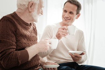 Talks and stories. Interested attentive handsome man sitting opposite his father holding a cup and listening.