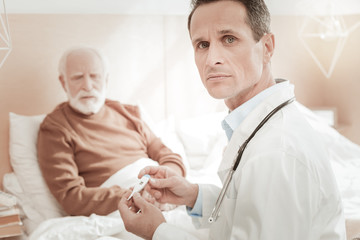 Fototapeta premium Bed news. Anxious skilled occupied medic sitting near his patient turning head and holding the thermometer.