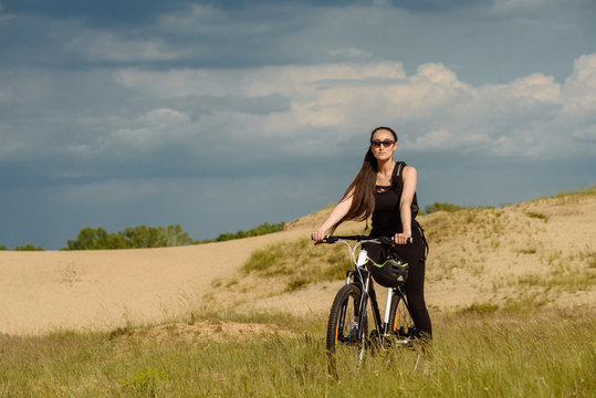 Sport and recreation. Full length image of young female cyclist. Young woman cyclist with backpack rides mountain bicycle on grass field on grey cloud sky