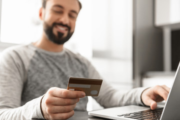 Image of satisfied man 30s in casual wear sitting at table in living room, and using laptop with credit card for making purchases online