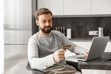 Photo of smiling man 30s in casual wear sitting alone in apartment, and using laptop with credit...