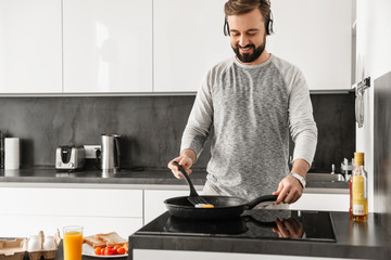 Cheerful man 30s wearing casual clothing frying eggs for breakfast, and listening to music via...