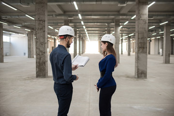 Two engineers, a man and a woman in construction helmets looking at a construction project. Construction site. The project is in the hands of a man. Business clothes. Concrete poles.