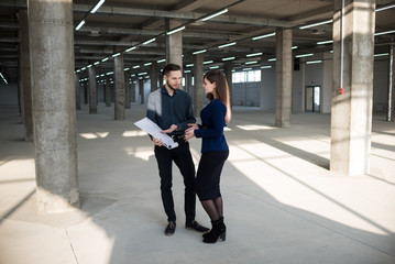 Two engineer-architects, a man and a woman discuss a construction project at a construction site. The project is in the hands of a man. TThe man explains and shows the plan. Business clothes. 