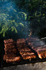 Grilled minced meat rolls called Mici or Mititei in traditional Romanian cuisine cooked outside on a hot summer day on barbeque grill with green small pine trees in background