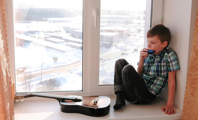 Playing a musical instrument. Boy plays the mouth organ sitting on the windowsill.