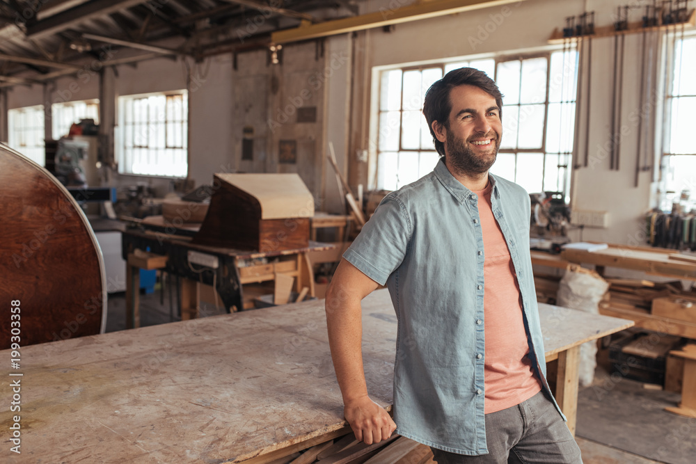 Wall mural Smiling young woodworker leaning on a table in his workshop