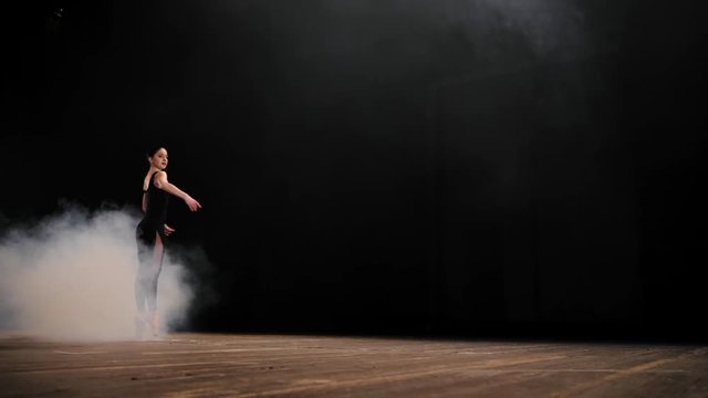 Young beautiful ballerina on smoke stage dancing modern ballet, spinning around diagonally. Classic movement Pique. Woman in black costume performs on scene. Slow motion.