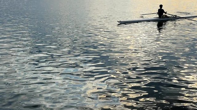 Slow Motion. Silhouette of young rower rowing on the tranquil sunset sea with the ripples on the water surface