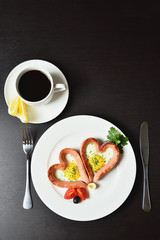 Scrambled eggs in the form of heart on a white plate with sausages, tomatoes, greens and coffee with lemon.