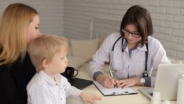 Woman doctor talking to the mother of a young boy and makes notes in a notebook, they sit in the office at the table. Pediatrics. Family doctor.