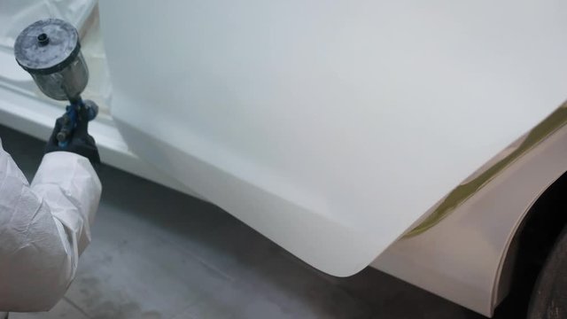painter is covering automobiles door in white color, sitting down and using industrial spray gun, close-up