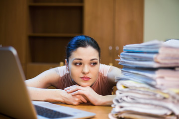 A young woman sits in front of a pile of papers and a computer holding her head