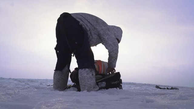 A skydiver in winter clothes is standing on the top of the hill and is wrapping his parachute and professional equipment in backpack. Purple winter sunset background