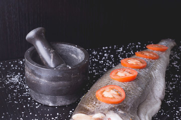 White fish with tomatoes and marble mortar on a black wooden background. Process of cooking