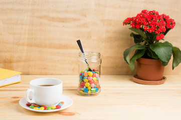 Coffee mug, red flowers, colored candy in a jar and a yellow diary on a wooden background. breakfast or lunch