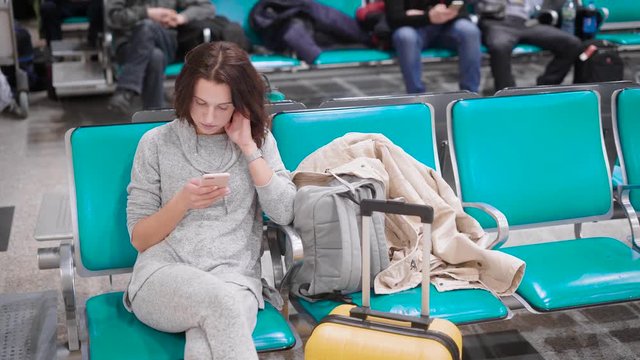 alone young woman is sitting on a chair in departure lounge of airport before flight, reading messages in her mobile